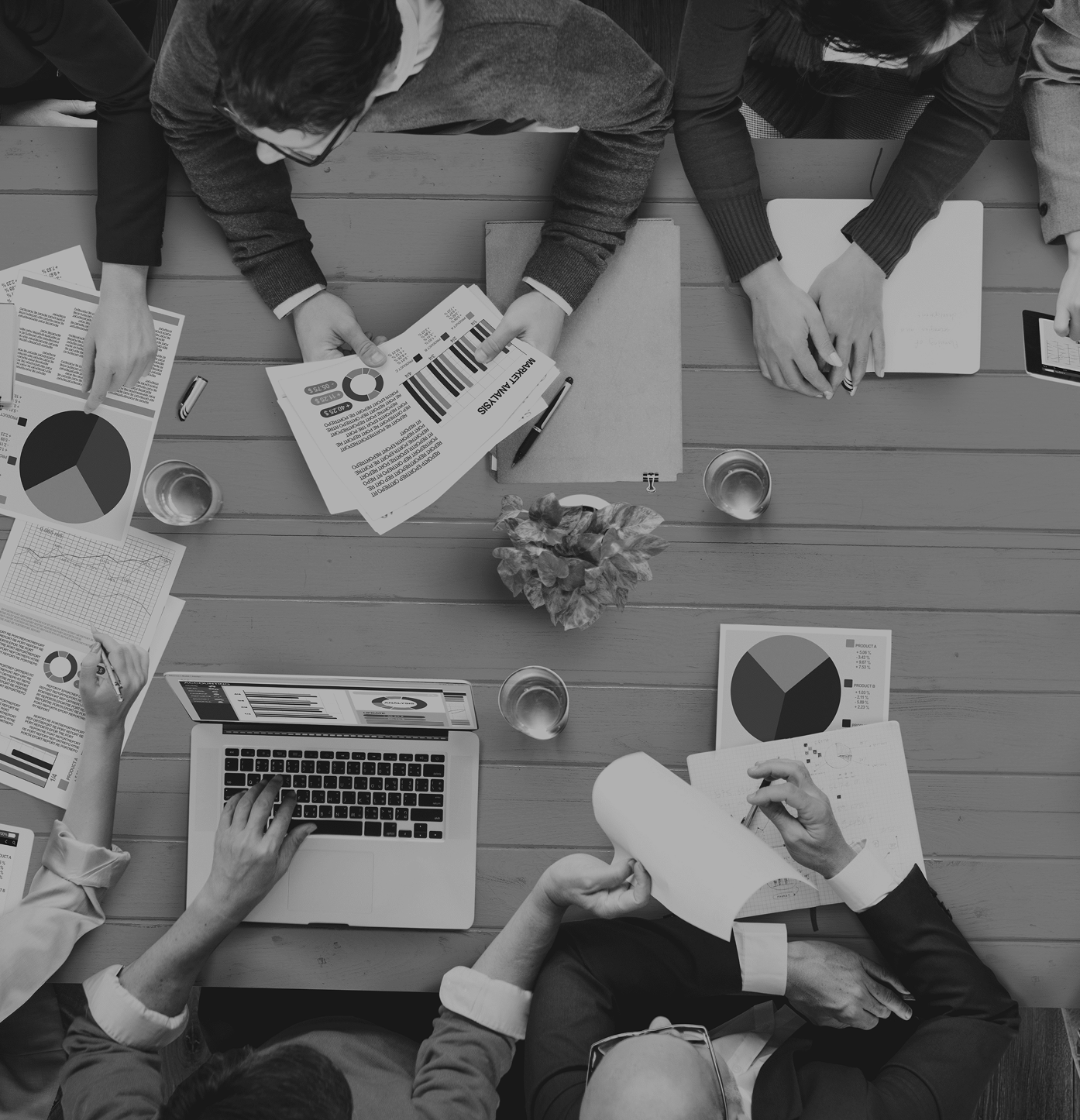 Overhead view of a business meeting with multiple people at a wooden table, analyzing charts and graphs, using laptops, tablets, and printed reports, with glasses of water and a small plant in the center.